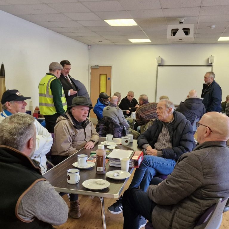 A group of older men sitting at tables in a communal room, drinking tea and chatting.
